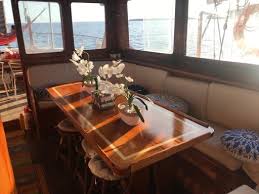 Caicco Interior of a boat with a wooden table, flowers, and seating by large windows.