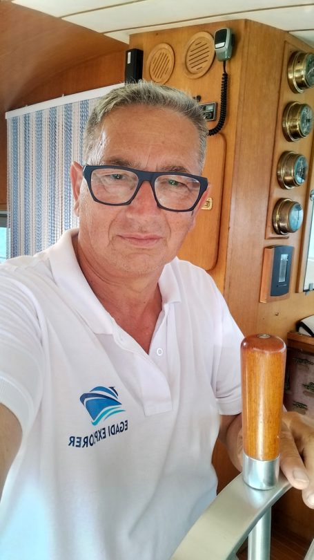 Comandante e Armatore "Pino" Man in a white polo shirt, wearing glasses, posing on a boat with nautical equipment.