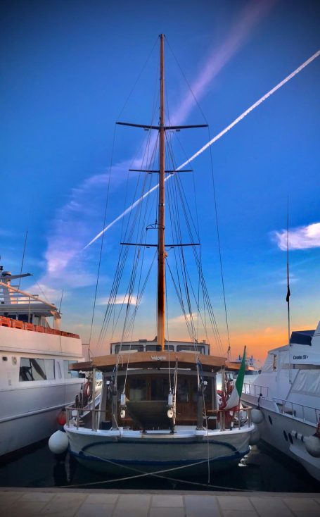 Caicco Sailboat docked between yachts against a vibrant sunset sky.